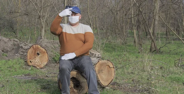 Bahar ormanında koruyucu maskeli ve eldivenli bir adam cep telefonuyla kendini fotoğraflıyor. Kavram adamı hiçbir yerde koronavirüs salgınından kendini güvende hissetmiyor.   