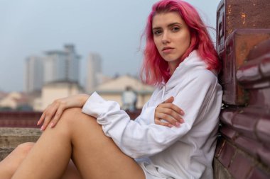 Çatıda kapüşonlu beyaz kazaklı pembe saçlı bir kızın portresi. 