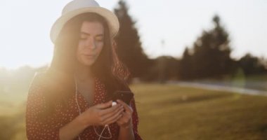 Elinde telefon olan kırmızı elbiseli mutlu bir kız gün batımında parkta müzik dinler. Telefonu ve kulaklığı olan gülümseyen genç bir kadın. Güzel kız müziğin tadını çıkarırken nazikçe gözlerini kapatır. Açık hava eğlencesi. 