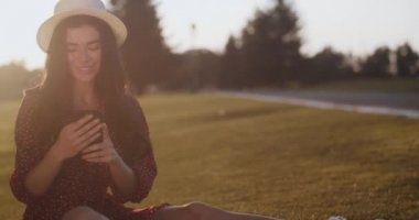  Gün batımında çimlerin üzerinde telefonla el sallayan sevimli kız. Çerçeveye bakıp güneşe gülümseyen güzel bir kadın. Güzel kız piknikte elinde telefonla oturuyor. Açık hava eğlencesi. 