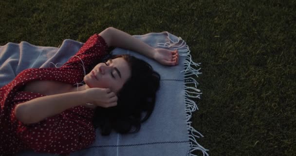 在夏天的公园里 穿着红衣服的漂亮的快乐姑娘在日落时听音乐 一个拿着电话和耳机的年轻女人可爱的女孩躺在草地上欣赏音乐 户外休闲 — 图库视频影像