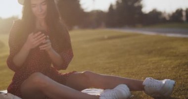 Şirin kız gün batımında çimlerin üzerinde oturan telefona bir mesaj yazıyor. Güzel bir kadın kadraja bakar ve ağır çekimde gülümser. Parkta piknikte telefonuyla oturan çekici bir kadın. Açık hava eğlencesi 
