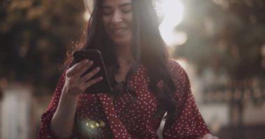 Parkta gün batımında elinde telefonla gülen mutlu bir kız. Yavaş çekimde güneşin altında dururken telefona bakan sevimli bir kadın. Elbiseli genç bir kadın sokakta telefonu kullanıyor.