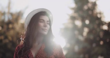 Gün batımında ağır çekimde poz veren hayalperest kız. Genç bir kadın yaz güneşinin altında parkta dururken nazikçe çerçeveye bakar.