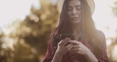 Elbiseli güzel kız gün batımında parkta bir mesaj yazıyor. Yavaş çekimde akıllı telefonlu tatlı bir kadın.