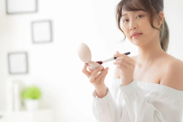 Beauty portrait young asian woman smiling with face looking mirror applying makeup with brush cheek in the bedroom, beautiful girl holding blusher facial, skin care and cosmetic concept.