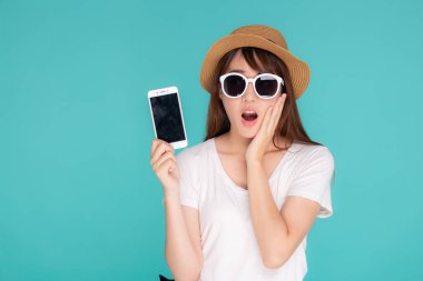 Güzel, genç Asyalı bir kadın tatilde sürpriz bir şekilde cep telefonunu sunuyor ve gösteriyor. Mavi arka plan, yaz gezisinde akıllı telefondan heyecanlanan bir kız, iletişim konsepti..