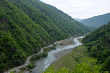 Dağ yolu, bir dağ nehri boyunca yeşil bir vadinin dibinde geniş açılı bir manzara esiyor.