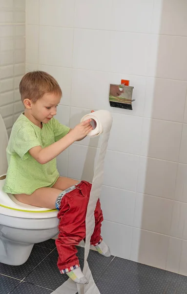 Το αγόρι κάθεται στην τουαλέτα στο δωμάτιο της τουαλέτας και ξετυλίγει ένα ρολό χαρτί υγείας Royalty Free Φωτογραφίες Αρχείου