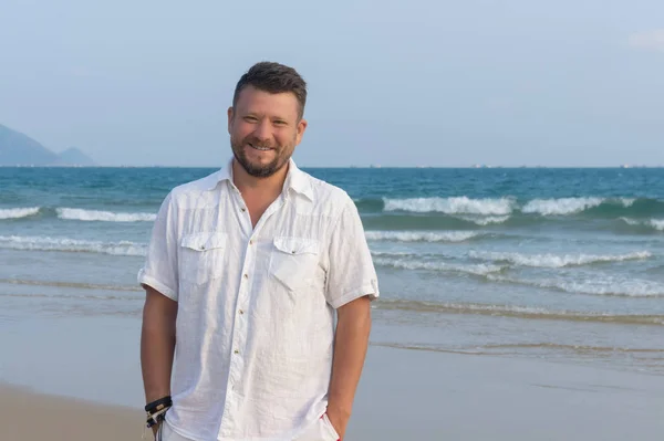 Portrait of a man with a middle beard in a white shirt, on the beach, on the background of the sea