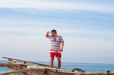 Geleneksel bir balıkçı teknesinde orta sakallı ve kırmızı şortlu bir adamın portresi güneşlik olarak gözlerini güneşten korumak için elini alnına koyup denize bakıyor..