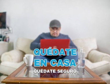 man an armchair with laptop in home office concept accompanied by coffee and antibacterial gel to avoid covid-19 or coronavirus. the photo has the phrase in Spanish stay home, stay save . quedate en casa quedate seguro clipart