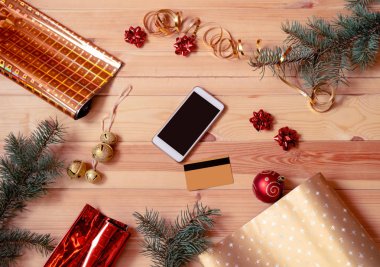 Akıllı telefon, kredi kartı, köknar dalı, ambalaj kağıdı ruloları ve tahta arka planda noel süslemeleri..