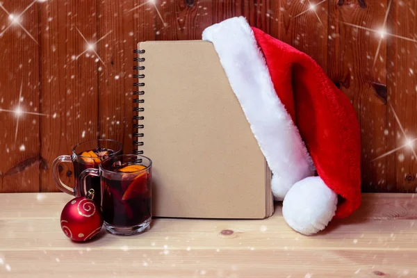 Prázdný zápisník, nadýchaný Santa klobouk, vánoční ples a svařené víno na dřevěném stole. — Stock fotografie