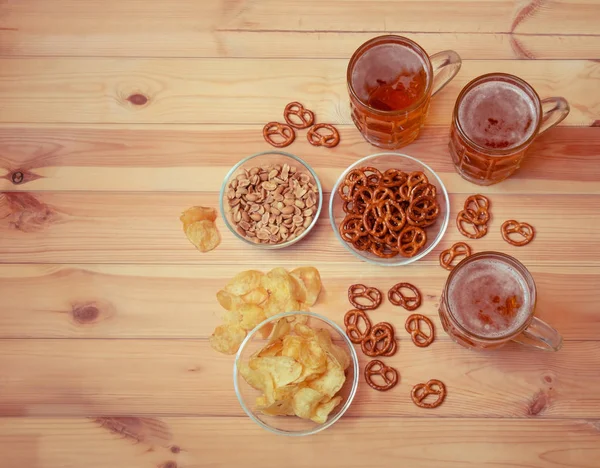 Tazas de cerveza ligera con papas fritas, pretzels salados y cacahuetes en la mesa de madera . — Foto de Stock