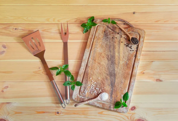 Leeres Holzschneidebrett, Gabel für Fleisch, Spachtel, Gewürze und grüne Basilikumblätter auf rustikalem Holzgrund. — Stockfoto