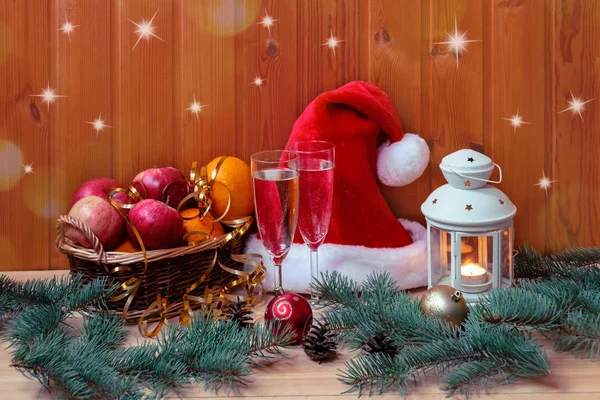 Wicker basket with fresh fruits, fluffy Santa hat, burning lantern, glasses of champagne and christmas balls. — Stock Photo, Image