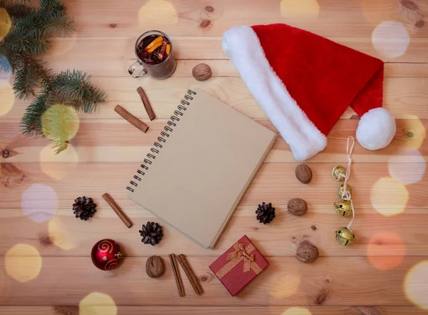 Prázdný zápisník, jedle, flétna Santa klobouk, vánoční ozdoby, sklenice svařeného vína a červené dárkové krabice na dřevěném pozadí. — Stock fotografie