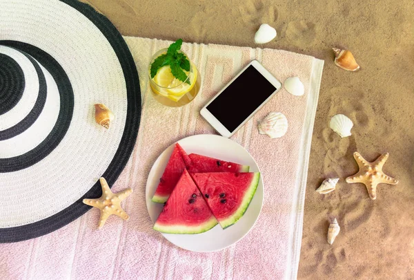 Grande cappello da spiaggia, smartphone, bicchiere di limonata e piatto di anguria su asciugamano sulla spiaggia di sabbia . Immagine Stock