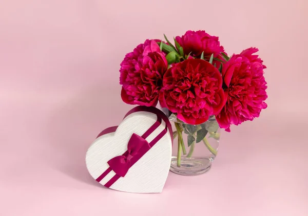 Rode pioenrozen in glazen vaas en hartvorm geschenkdoos op roze achtergrond. Selectieve focus. — Stockfoto