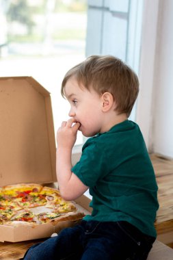 Küçük çocuk pencere kenarında oturur ve bir kutudan pizza yer..