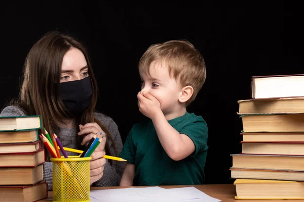 Mom in a medical mask sits near her son at a desk with books and pencils. the child covers his mouth with his hand. quarantined distance learning. family leisure.