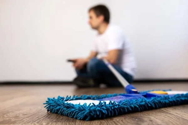 a young man sits on the floor against the background of a mop with a phone in his hands. rest during the general cleaning. the guy is out of focus.