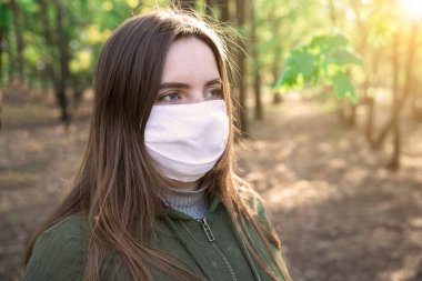Ağaçların arkasında, parkta maske takan genç bir kız. Yakın plan portre. Virüslere ve enfeksiyonlara karşı koruma. Coronavirus 'a karşı savaş. Karantina yürüyüşleri. Bakışlar.