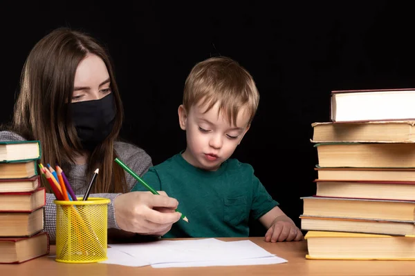 Mom in a medical mask helps her son learn to write. at a table with books on a black background. quarantined distance learning. difficulties of online lessons.