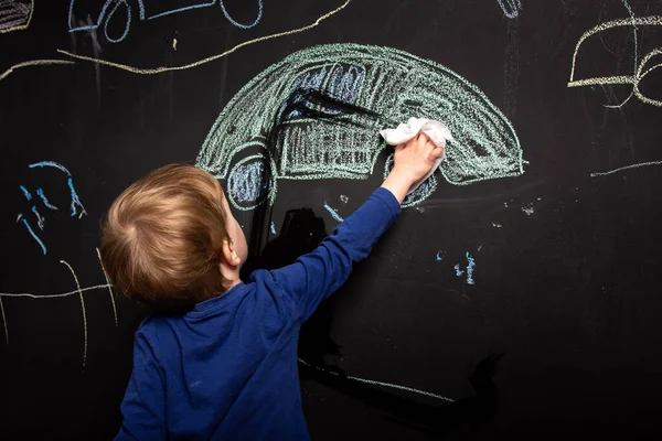 little boy erases the drawing from the board. chalk drawn cars on black background. children\'s creativity.