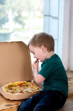 Küçük çocuk pencerenin yanında oturur ve kutudan bir dilim pizza yer. Pencereden sokak görüntüsü