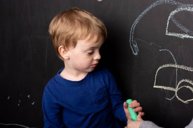 Küçük çocuk siyah tebeşirli siyah bir tahta duvarın yanında duruyor. Karantinaya alınmış ev eğlencesi. Arka planda çocukların çizimleri var.