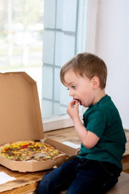 Apartmanın penceresinin yanında pizza yiyen küçük aç bir çocuk. Çocuk ellerini ağzına bir domates dilimi sokuyor. yakın plan.