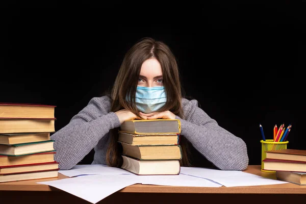 a young girl in a medical mask at the table laid her head on books. the difficulties of distance learning in quarantine. infection protection.