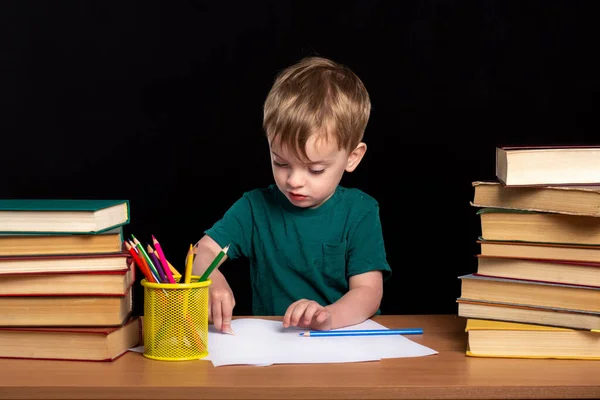 a small child draws with a pencil at a desk next to two stacks of books. Black background. isolate. classes at home.