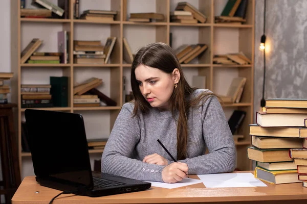 a young girl sits at a table with a laptop and writes out information in documents. stacks of books near and a bookcase on the background. difficulties of remote work. office weekdays. the teacher is preparing for the lesson. freelancer