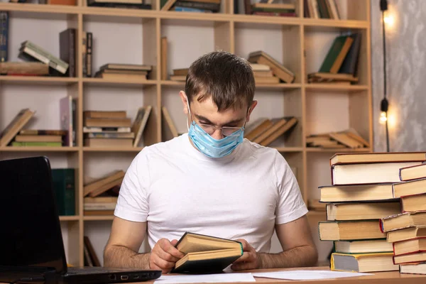 young man in a medical mask sits at a table with laptops and reads a book. Difficulties of online learning in quarantine. remote office work. bookcase on the background.