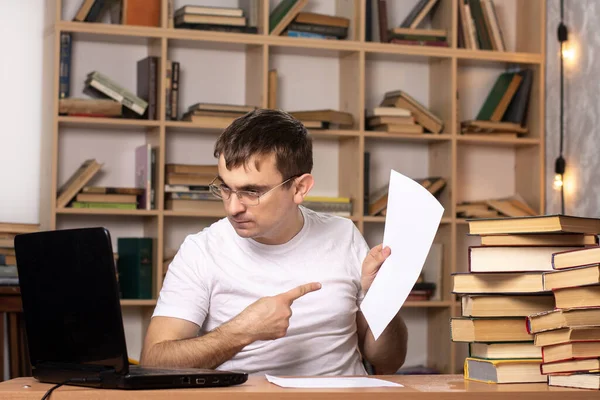 白いTシャツを着た若い男がノートパソコンを持ったテーブルに座り 書類を指し示している 本の山と背景の本棚です 遠隔作業の難しさです 平日営業 — ストック写真