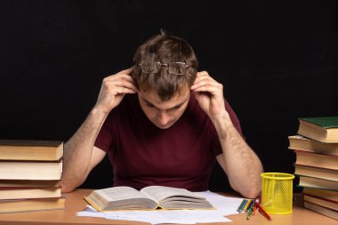 Bir öğrenci masada oturur ve kafasını elinde tutan bir kitap okur. Siyah arka plan. Genç adam sınava hazırlanıyor. Mesafe öğrenme zorluğu. İş için serbest çalışan. Metin için yer