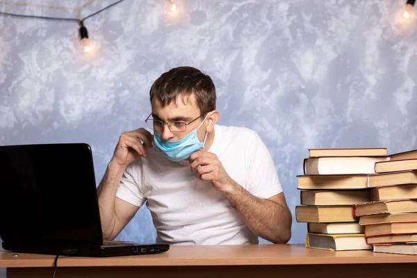 young man in a medical mask at the desk with books and laptop. Angry office worker. Quarantined emotions.