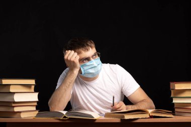 Genç bir adam elinde kalemle ve düşünceli bir bakışla tıbbi maskeli kitaplarla masada oturuyor. Karantina zorlukları. Siyah arka plan. Bir salgın sırasında enfeksiyonlara karşı koruma.