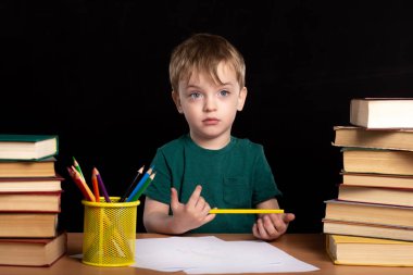 Masada elinde kalemle oturan üç yaşındaki bir çocuk iki kitap yığınının yanında oturuyor. Bir yana bak. Siyah arka plan. İzole et. Ödev dersleri. uzaklık öğrenme