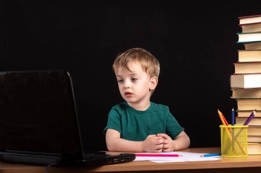 Küçük çocuk, kitaplarla ve kalemlerle dolu bir masada dizüstü bilgisayardan ders izliyor. Siyah arka plan. Erken kalkınmada ciddi görün. uzak eğitim çocuğu