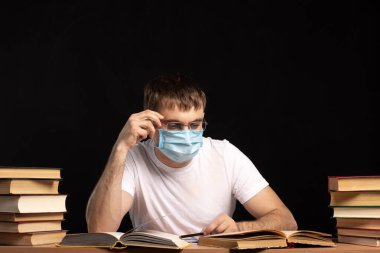 Masadaki tıbbi maskeli genç adam siyah bir duvara karşı kitap okuyor. Öğrenci karantinada. Uzaktan öğrenme. ofis çalışanı