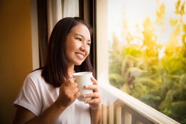 Азиатка держит чашку кофе расслабляющий взгляд за окном — стоковое фото