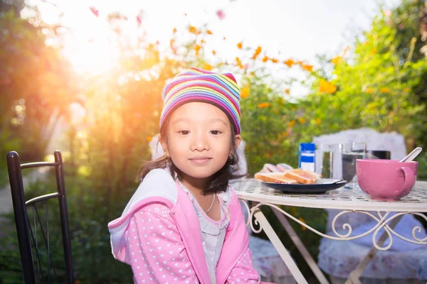 Liten asiatisk tjej njuta av att äta bröd till frukost i trädgården — Stockfoto