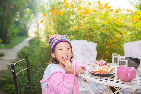Liten asiatisk tjej njuta av att äta bröd till frukost i trädgården — Stockfoto