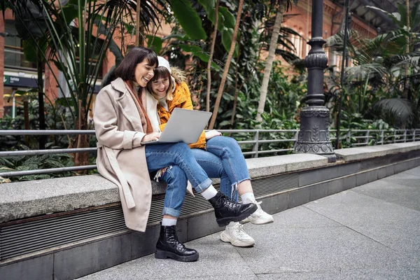 Stock photo of a side view of two caucasian girls using a laptop. They are seated on a stone bench inside a train station.