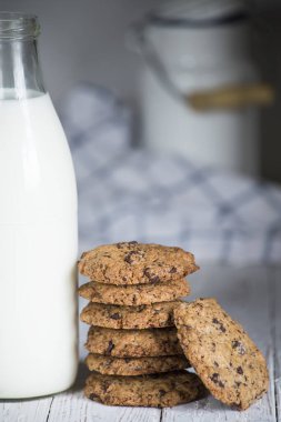 Süt şişesinin yanına istiflenmiş çikolatalı kurabiye beyaz tahta bir tahtanın üstünde, arka planda süt sürahisi var.