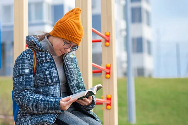 Clever teen girl is reading a book in the playground. Children reading, learning in the park, student girl